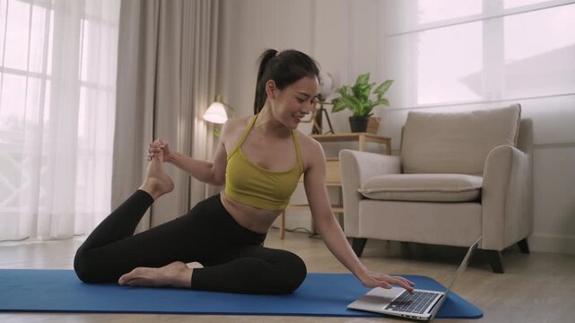 Happy Asian young woman yoga instructor teaching, talking, and video chatting on laptop webcam camera in plank position on a mat for healthcare in living room at home. Fitness training class concept. 