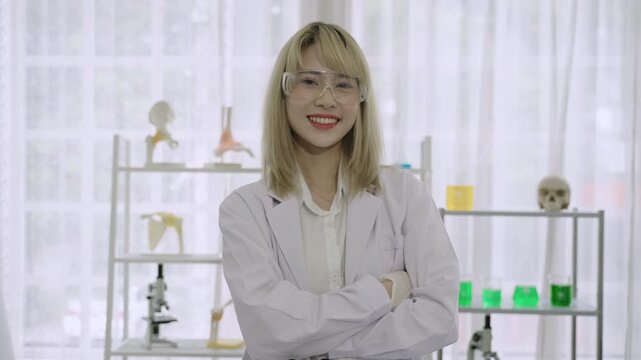 Portrait of happy beautiful Asian young woman scientist wearing a white lab coat and safety goggles standing smiling with arms crossed and thumb up while looking at the camera in a science laboratory.