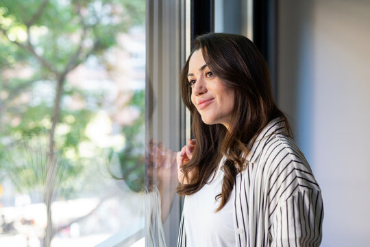 A young woman stands by a large window, gazing outside with a thoughtful, gentle smile. Soft natural light and a calm home setting create a mood of reflection, hope and peaceful relaxation.