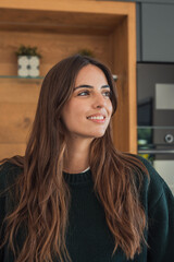 Obraz premium Smiling young woman looks thoughtfully to the side in a cozy home kitchen portrait. Natural light highlights her long hair and relaxed expression, conveying warmth, confidence and everyday