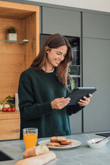 A young woman stands at a kitchen island smiling as she reads a tablet during a relaxed morning breakfast. Toast, bread and a glass of orange juice sit on the counter nearby. © Daniel