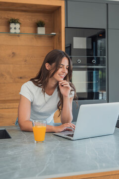 A young woman leans on a kitchen island, smiling as she works on a laptop with a glass of orange juice nearby, conveying relaxed remote work, productivity and casual home lifestyle.