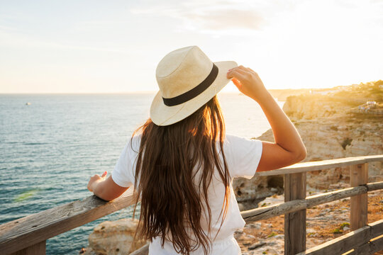 Woman wearing a hat, enjoying a peaceful moment while looking at the sunset over the ocean from a cliffside viewpoint during her summer holidays