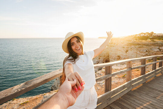 A smiling young woman in a sun hat holds her partner's hand and leads them along a wooden boardwalk by the sea at golden hour, conveying travel, romance and joyful summer vibes.