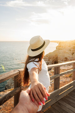 Woman wearing a straw hat and white outfit, holding hands with a partner, exploring a wooden walkway along the cliffs overlooking the ocean during a golden sunset, representing travel and adventure
