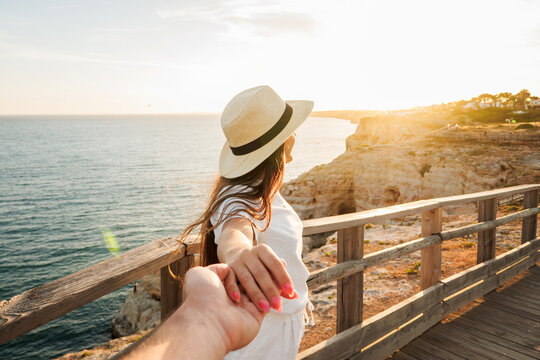 Romantic travel moment of a couple holding hands on a wooden boardwalk overlooking rocky cliffs and the ocean at sunset, evoking adventure, connection, summer vacation and wanderlust.