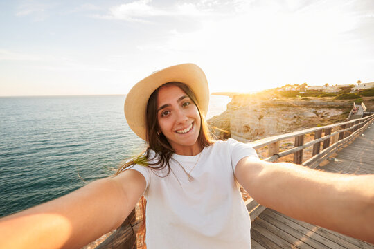 Young woman wearing a hat smiling at camera, taking a selfie on a wooden boardwalk overlooking the ocean and coastline during a golden sunset, symbolizing happiness and freedom while traveling
