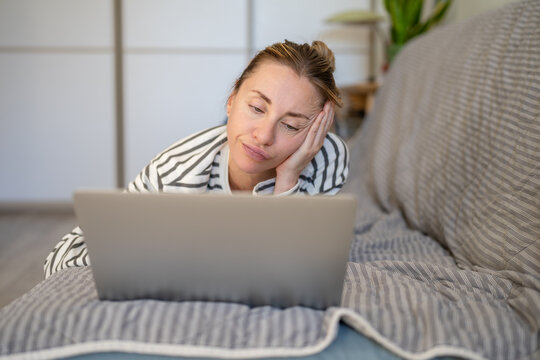 Bored uninterested woman browsing online on laptop watching media while lying on couch at home, lazy female relaxing on sofa with displeased expression surfing on computer, wasting time online.