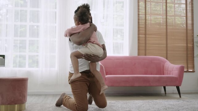 A cute African American child daughter is running and smiling to embrace her father while sitting and waiting on the carpet floor, happy and bonding in the living room at home. Father's Day concept. 