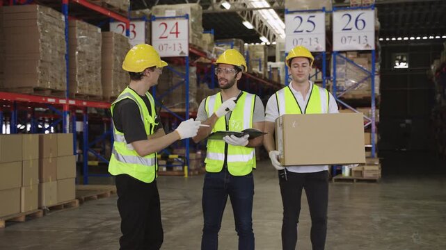 Teamwork of three warehouse workers in uniform wearing yellow safety helmets, standing and working talking and discussing planning in large logistics distribution factory. Inventory stock concept. 