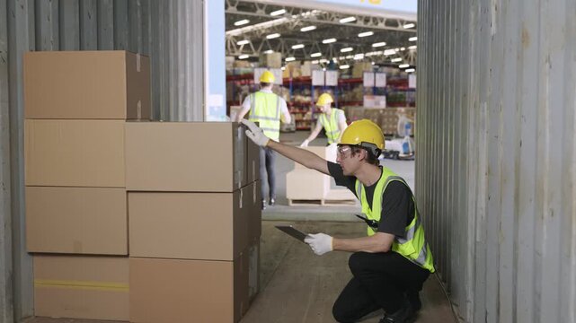 Team of young men warehouse workers in uniform wearing yellow safety helmets helping, holding, and carrying cargo cardboard boxes for unloading from a container in a logistics distribution factory. 