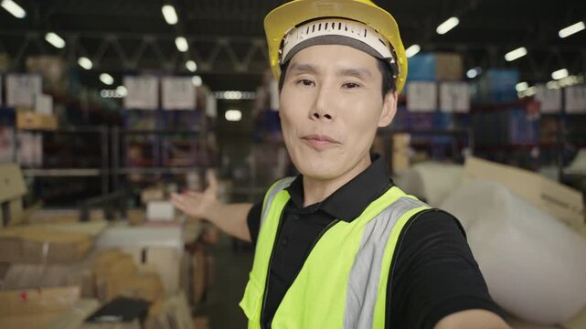 Happy, confident Asian man warehouse worker in uniform wearing yellow safety helmet, standing, smiling, and holding camera while talking and greeting via video call in logistics distribution factory. 