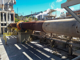 Naklejka premium Large industrial rotary kiln and heavy machinery infrastructure in a manufacturing plant under a blue sky.
