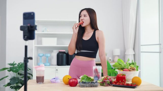 Asian digital creator sharing daily detox routine with organic fruits and blender. Active young woman live streaming fitness journey and nutritional advice from home kitchen.