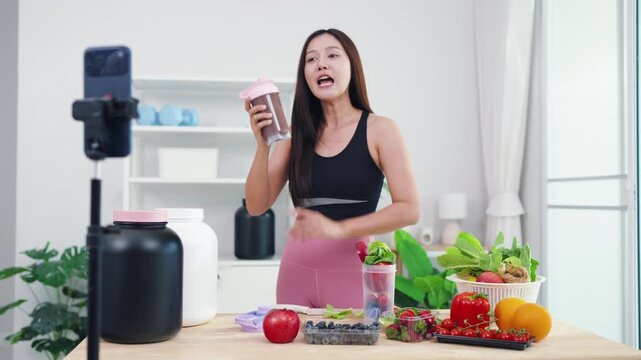 Asian digital creator sharing daily detox routine with organic fruits and blender. Active young woman live streaming fitness journey and nutritional advice from home kitchen.