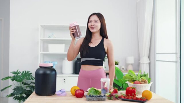 Asian digital creator sharing daily detox routine with organic fruits and blender. Active young woman live streaming fitness journey and nutritional advice from home kitchen.