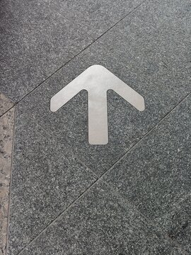 Metal directional arrow sign embedded in gray textured granite floor tiles pointing forward.