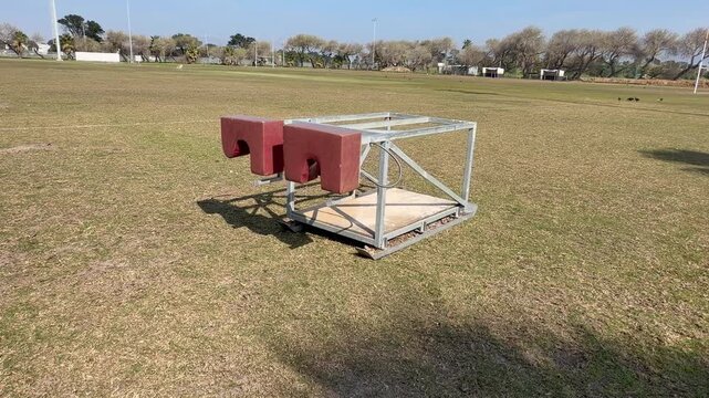 : A weathered metal rugby scrum machine with red padded sled sits idle on a grassy sports field under a clear sky, training equipment framed by distant trees and pavilion buildings.