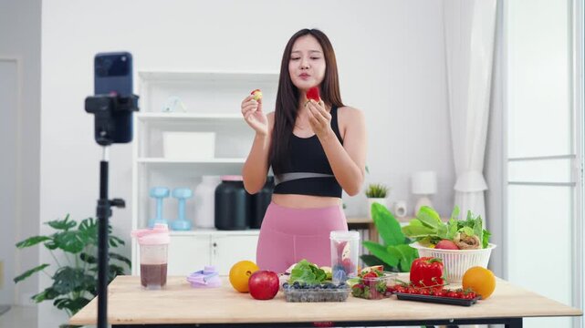 Asian digital creator sharing daily detox routine with organic fruits and blender. Active young woman live streaming fitness journey and nutritional advice from home kitchen.