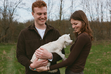 Naklejka premium Cheerful couple outdoors, smiling while cradling a goat kid, sunlit meadow scene, relaxed posture, natural colors, candid lifestyle moment, warm interaction in a peaceful field setting