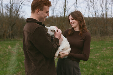 Naklejka premium Casual couple outdoors gently care for a white goat kid, sharing a tender moment in a rustic field, soft natural light, warm sweaters, calm smile, and peaceful, affectionate mood