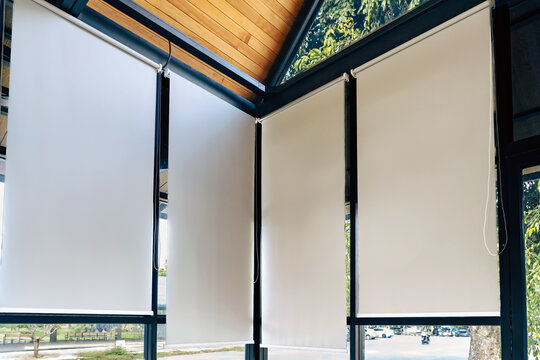 White roller blinds installed on large windows of modern room. 
Inside view of grey fabric roller blinds or shades installed on big glass windows providing sun protection in contemporary interior.