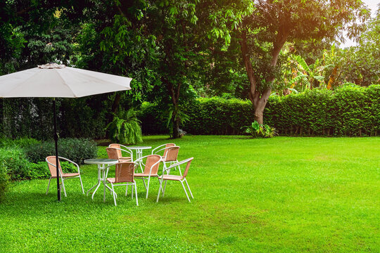 Garden patio set with white umbrella on lush green lawn. 
Outdoor seating area setup featuring woven wicker chairs, round tables, and large white patio parasol placed on vibrant green grass field.