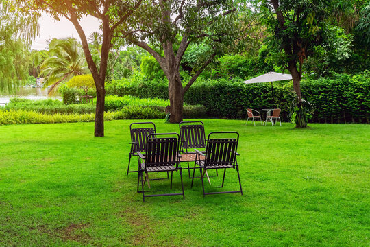 Outdoor patio seating sets on green lawn by river. Garden chairs and wooden tables on lush green grass field under shady trees in lakeside backyard, featuring peaceful riverfront landscape view.