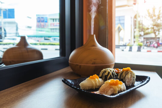 Sushi dish on window counter bar with ultrasonic humidifier. Assorted Japanese sushi, including nigiri and gunkan topped with roe next to wood grain aroma diffuser oil diffuser by window view.