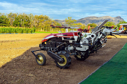 Agricultural tractor standing with front blade and rear rotary tiller on tilled agricultural field during land preparation for farming with trees and cornfield in background. Agricultural machinery.