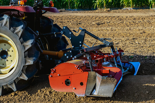Agricultural tractor standing with front blade and rear rotary tiller on tilled agricultural field during land preparation for farming with trees and cornfield in background. Agricultural machinery.
