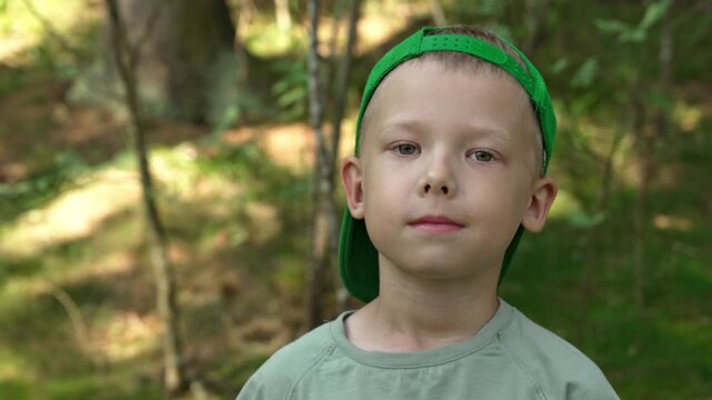 Cute little boy wearing a green baseball cap blinking and looking at the camera while standing in a beautiful sunlit forest, showcasing a calm and peaceful expression during a summer outing