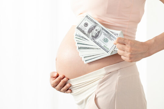Pregnant woman holding US dollar bills over belly to represent pregnancy costs