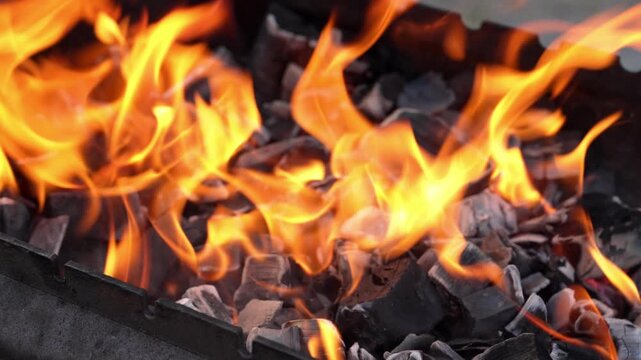 Golden tongues of fire flicker intensely above smoldering black coals in a metal grill under the warm afternoon sun as preparation begins for a delicious backyard feast with friends and family members