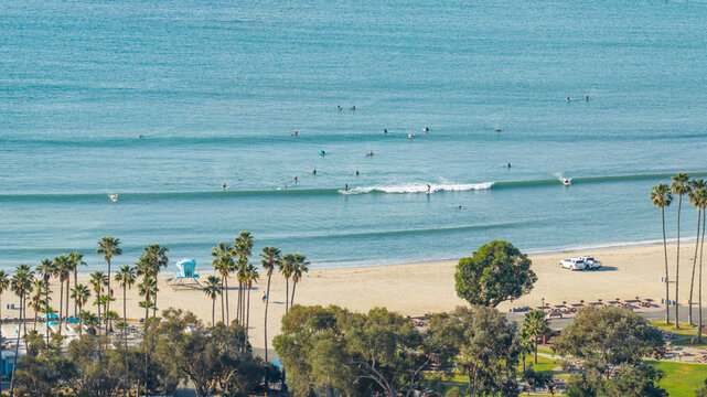 Aerial view of Dana Point beach with surfers riding waves in the Pacific Ocean, palm trees, a lifeguard tower, and vehicles parked on the sand in Dana Point, California, United States.