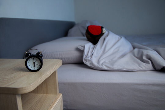 There is a black alarm clock on a wooden bedside table, and in the background a brown bear in a red mask is covered with a gray pastel blanket. The concept of early awakening, healthy sleep, sleep