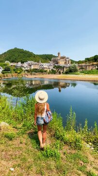 young female traveler with straw hat and backpack viewing estaing medieval village across river in aveyron france