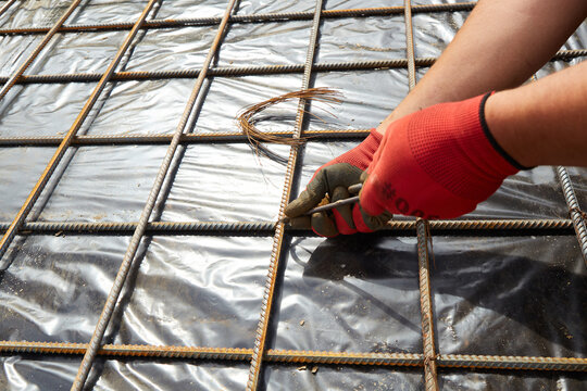 Construction worker ties steel reinforcement bars with binding wire over waterproof membrane before concrete pouring.