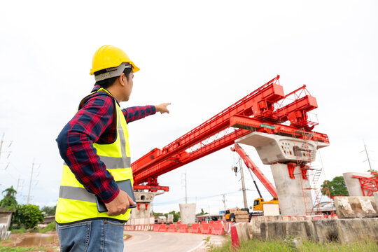 Asian engineer inspecting and pointing at a large bridge construction project with launching gantry, representing civil engineering, infrastructure development and construction safety.