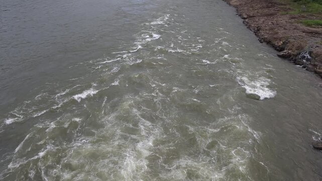 High angle overview of a widening trail of churning river rapids and white foam flowing rapidly downstream past a rocky bank, conceptualizing fluid dynamics, hydraulic pathways, & roaring soundscapes.