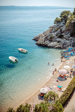 Aerial view of the beach in Brela with turquoise water, small boats, rocky cliffs, and people relaxing on the sand in Brela, Split-Dalmatia County, Croatia.