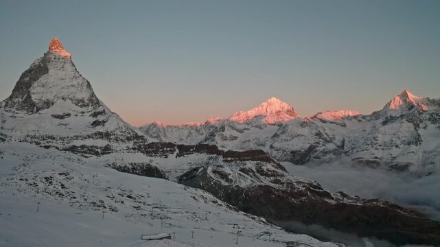 Sunrise panorama featuring the Matterhorn, Dent Blanche and the Zermatt ski resort glowing with alpenglow beneath a clear morning sky in the Swiss Alps.