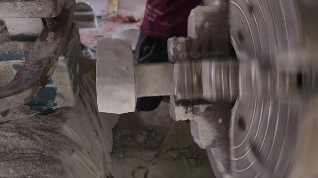 Slow-motion horizontal footage of an Indian artisan shaping a granite stone utensil on a rotating lathe machine. The spinning stone is carefully worked with cutting tools to create a rounded form