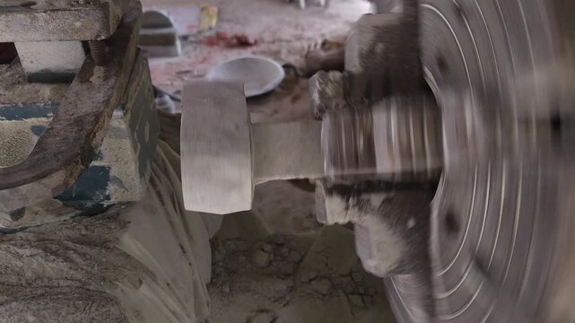 Slow-motion horizontal footage of an Indian artisan shaping a granite stone utensil on a rotating lathe machine. The spinning stone is carefully worked with cutting tools to create a rounded form
