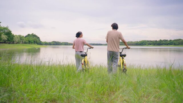 Rear view of young couple standing with bicycles on grass near a lake.