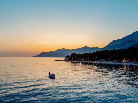 Aerial view of a small boat on the calm Adriatic Sea near the pine-covered coastline at sunset, with mountains in the background in Brela, Split-Dalmatia County, Croatia.