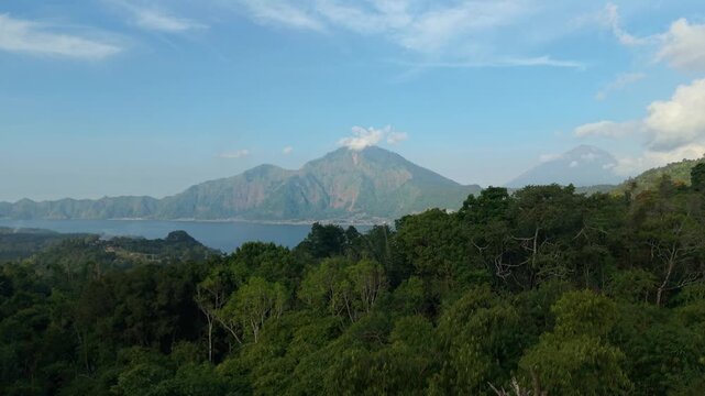Aerial view of Mount Abang rising above lush tropical forests and the waters of Lake Batur under a clear blue sky in Kintamani, Bali, Indonesia.