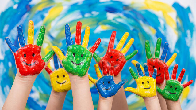 Many children's hands painted with vibrant colors and smiley faces are raised high together against a blue and yellow backdrop.
