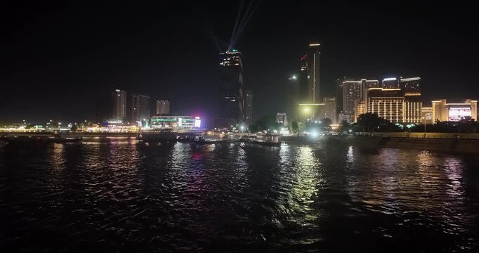 Night view of the Phnom Penh skyline, featuring searchlights from a skyscraper and ferries moored at the city's ferry terminal, as seen from a boat.