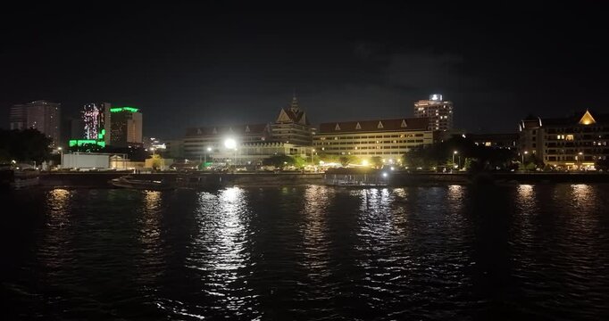 Panoramic night view of the Phnom Penh riverfront, showcasing luxury hotels and city skyscrapers casting shimmering reflections onto the dark, rippling waters of the Tonle Sap, as seen from a boat.